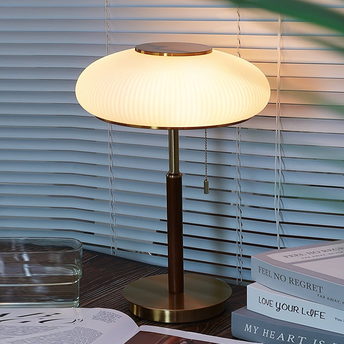 Table lamp on a surface with books and a glass container, blinds in the background