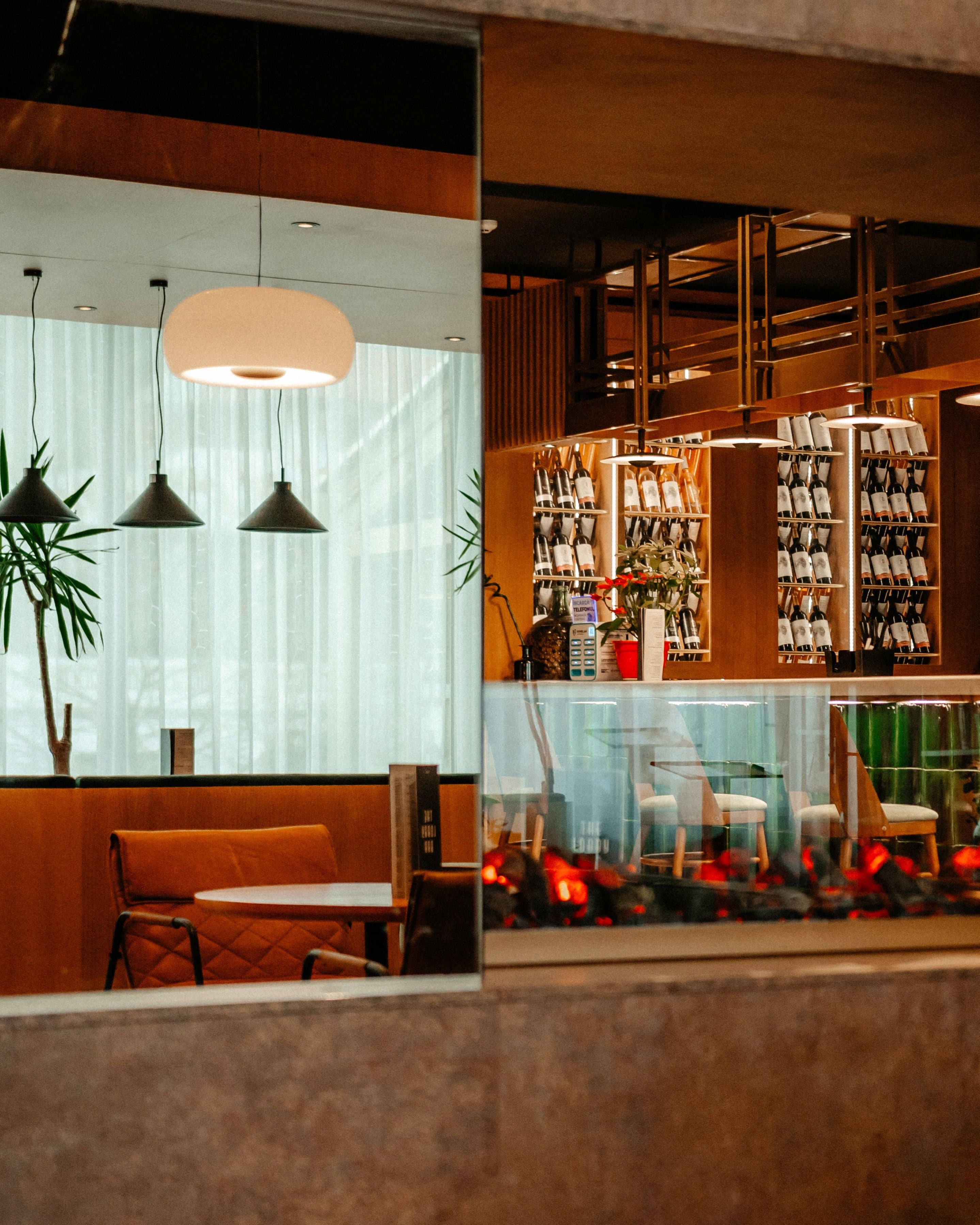 An interior architectural view of a modern bar and dining space with warm wood paneling, pendant lights, glass partitions, and a fire pit with glowing embers in the foreground.