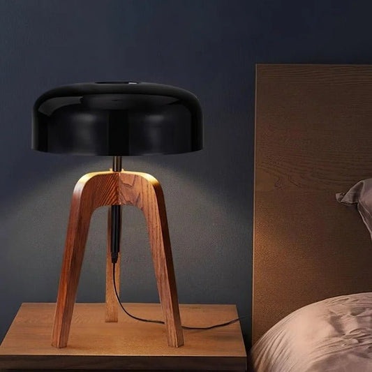 Table lamp with wooden base and black shade on a wooden surface against a dark wall.