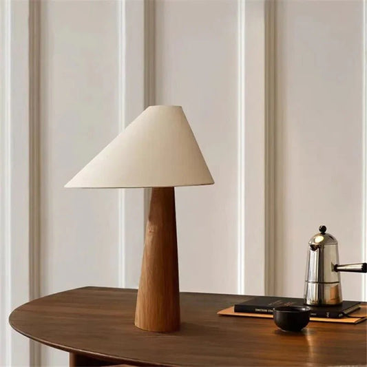 Table lamp with wooden base and beige shade on a wooden surface with a curtain background
